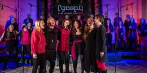 Eine Sängergruppe des Grazer Gospelchors glänzt auf der Bühne mit Mikrofonen, während ein harmonischer Chor für eine mitreißende Kulisse sorgt.